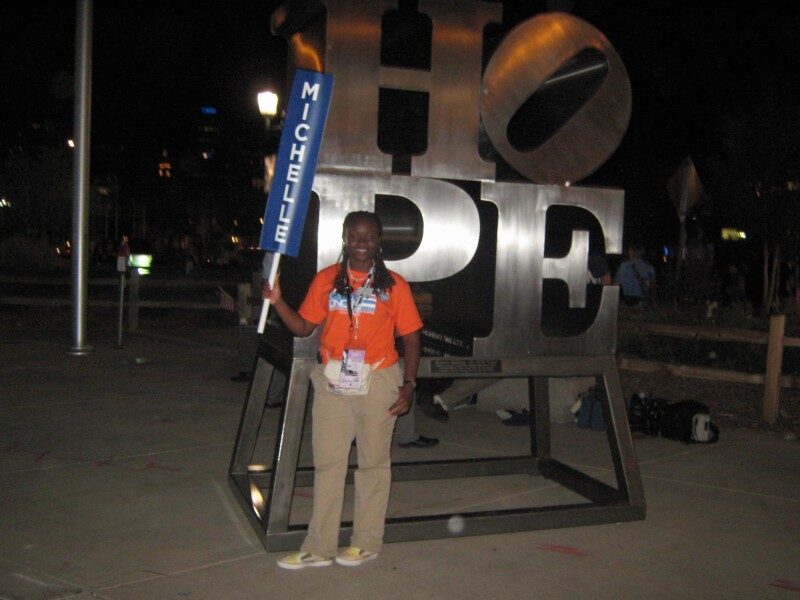 Sajdah holding a sign reading "MICHELLE" in front of a large illuminated HOPE sculpture, 2008