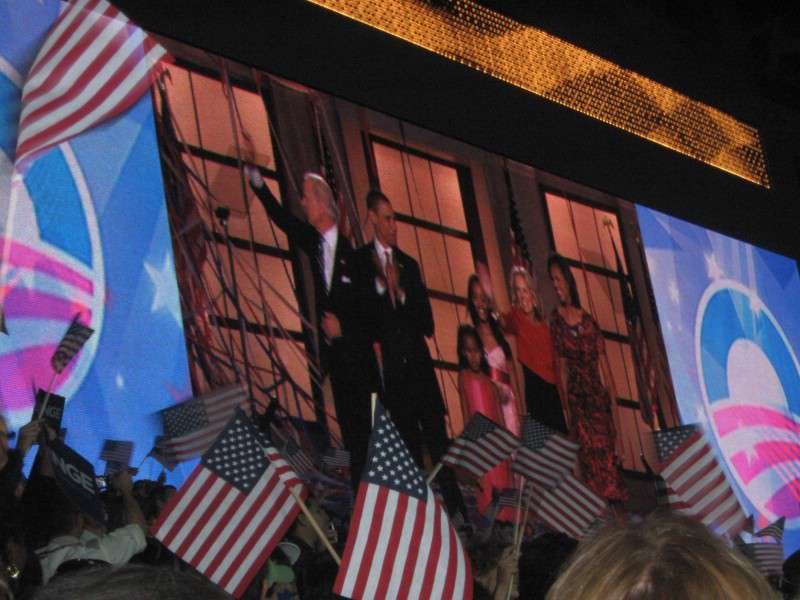 Large screen showing the Obama and Biden families waving on stage, flags in foreground