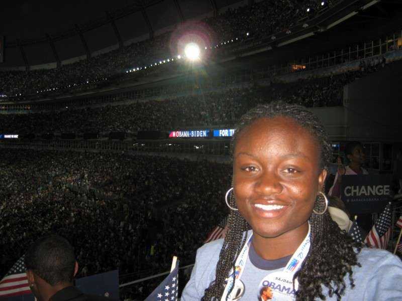 Sajdah smiling in a packed nighttime stadium, “OBAMA-BIDEN” visible on a screen behind her
