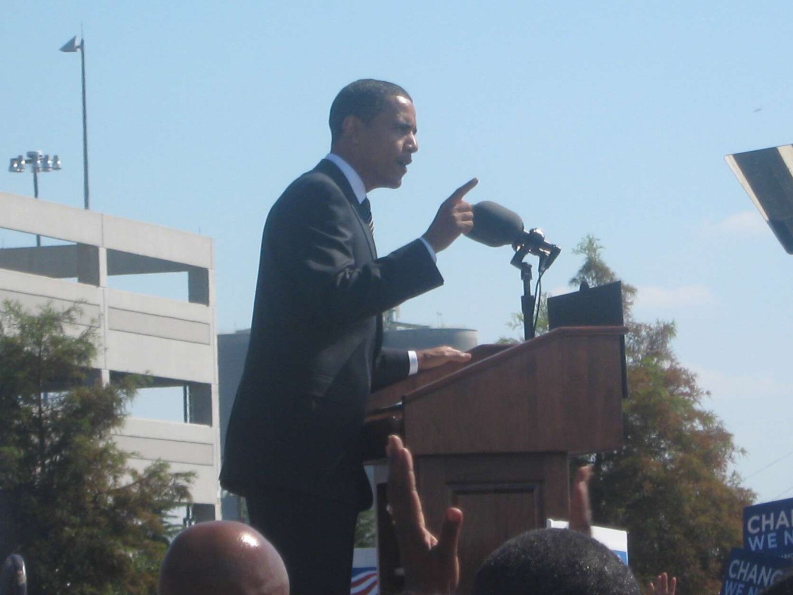 Barack Obama speaking at an outdoor podium, gesturing, with “Change We Need” signs in the crowd