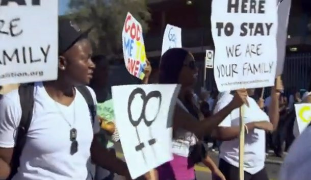 Sajdah marching in a crowd carrying marriage-equality signs reading "Here to Stay" and "We Are Your Family"